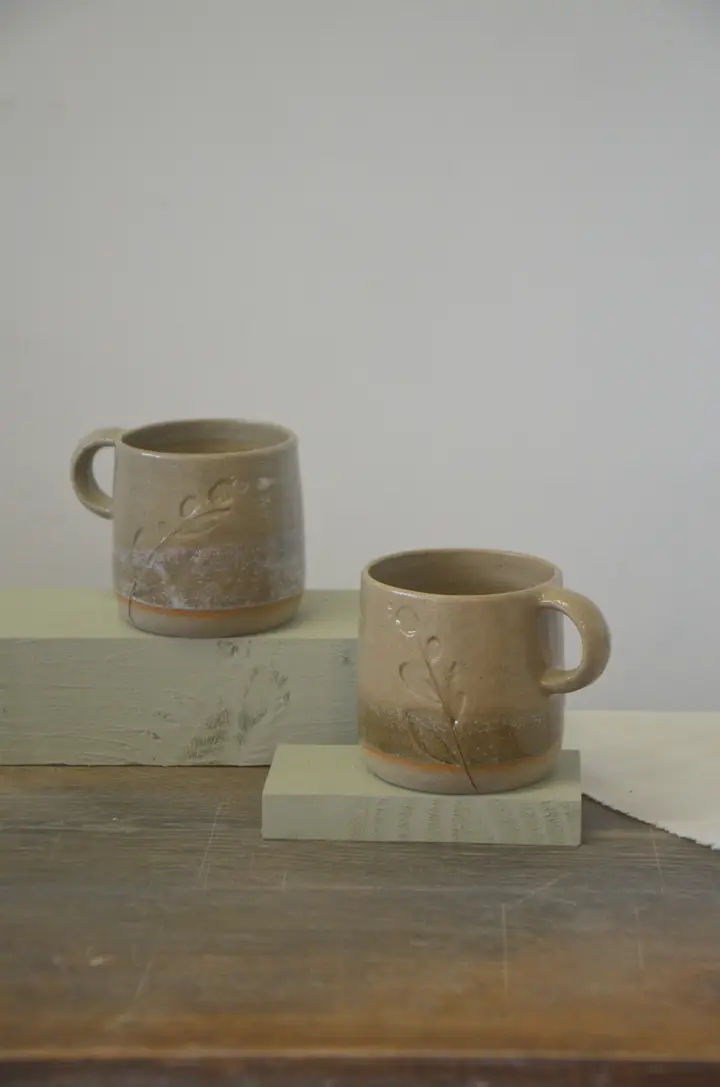 Deux tasses beiges à anse, décor de brins végétaux, posées sur des socles.