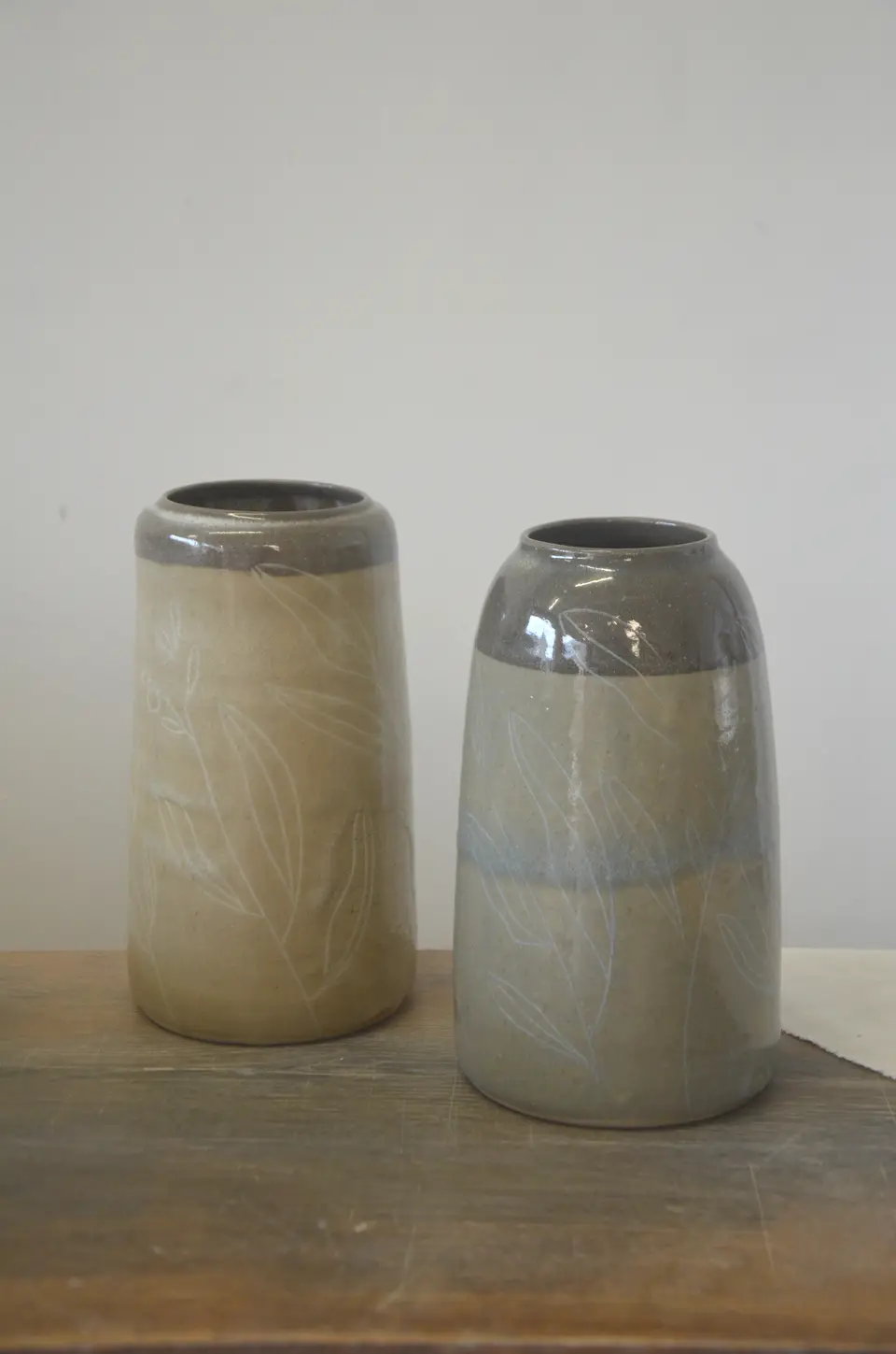 Deux vases à col gris, décor de feuillages clairs, posés sur une table en bois.