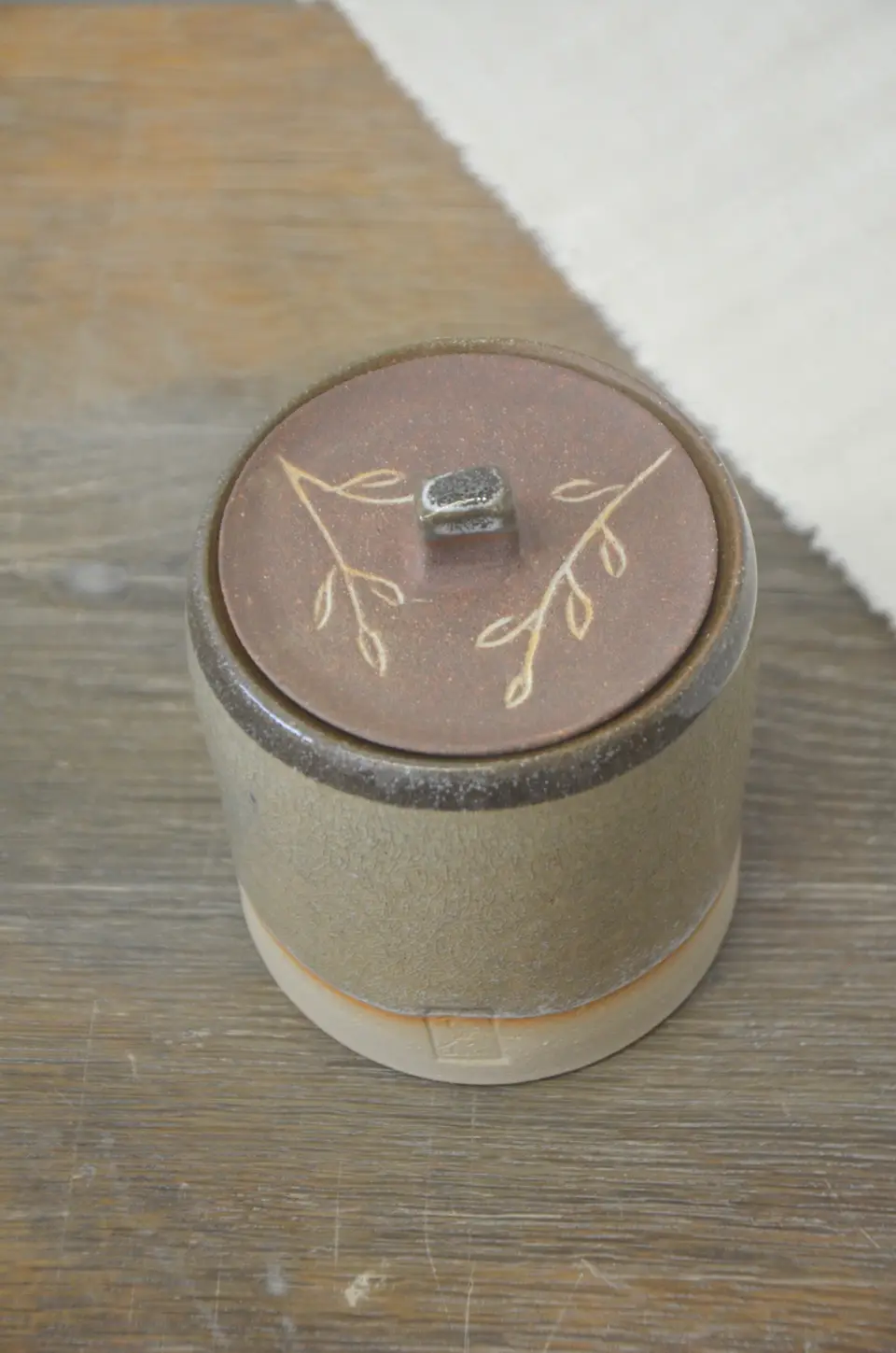 Pot à couvercle brun décoré de brins de feuillage, vu de dessus.