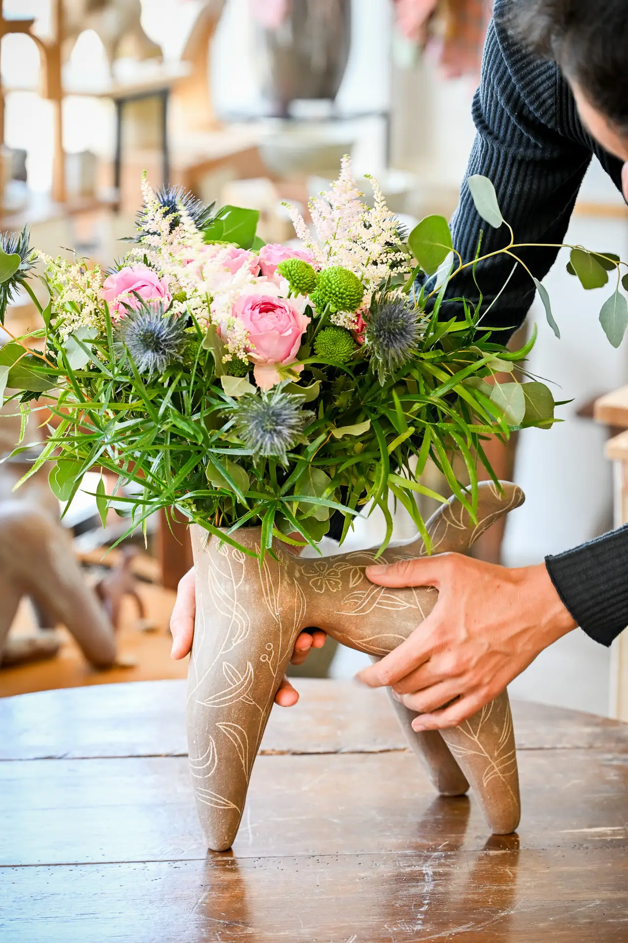 Deux mains posent sur une table une pièce brune à quatre pieds garnie d'un bouquet de fleurs.